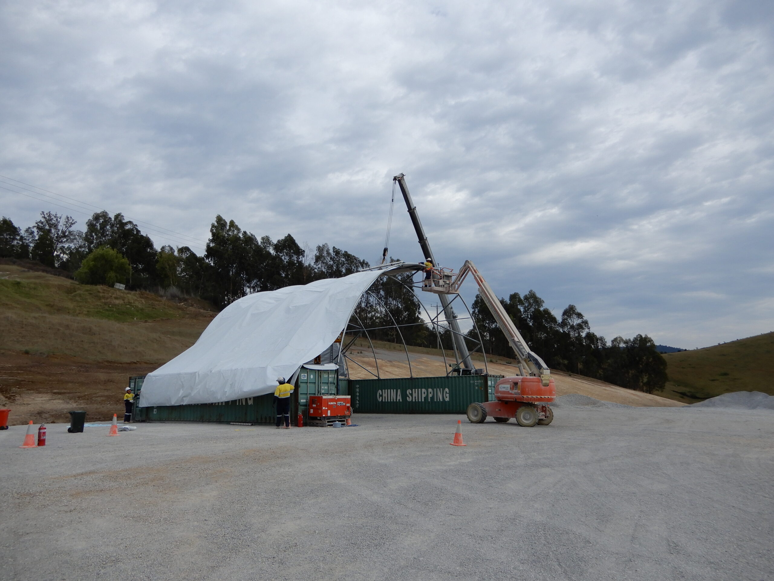 Workers assembling a container dome shelter on a construction site using heavy machinery and support frames.