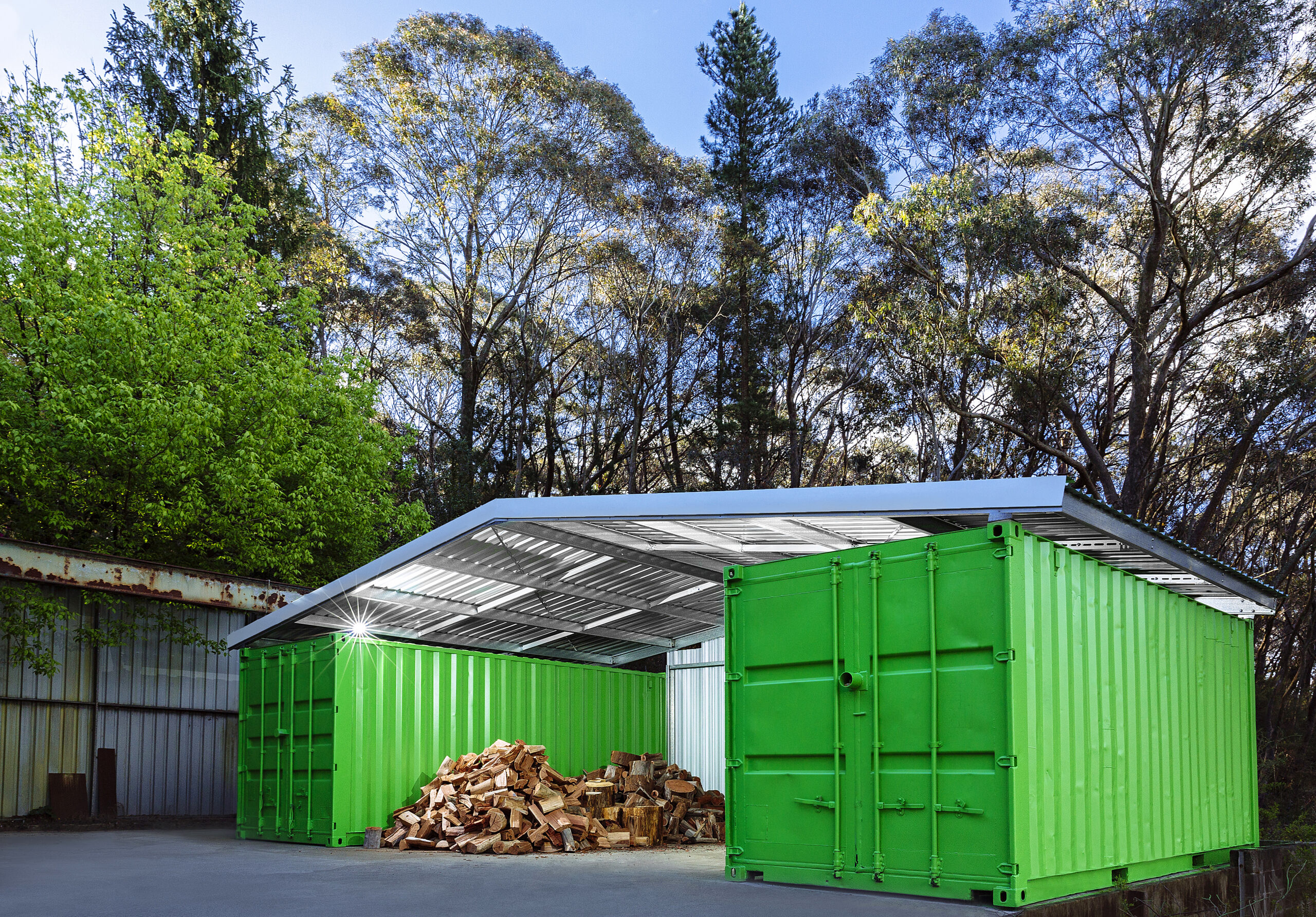 A shelter made of green shipping containers with a metal roof, storing wood logs near a forested area.